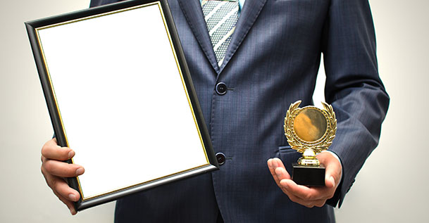 A closeup of a man in a suit holding a small trophy in one hand and an empty picture frame in the other.