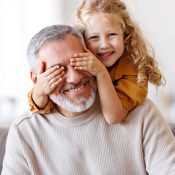 A young girl standing behind an elderly man with a beard covering his eyes with her hands.