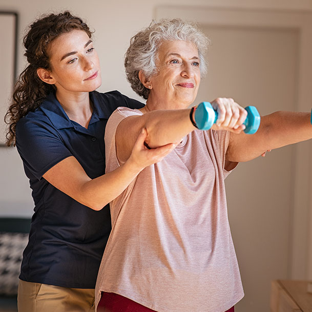 A young woman helping an elderly woman who is holding hand weights.