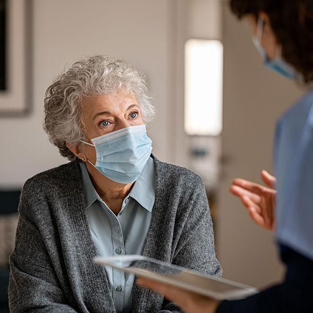 An elderly woman wearing a mask looking at a doctor wearing a mask and holding a tablet.