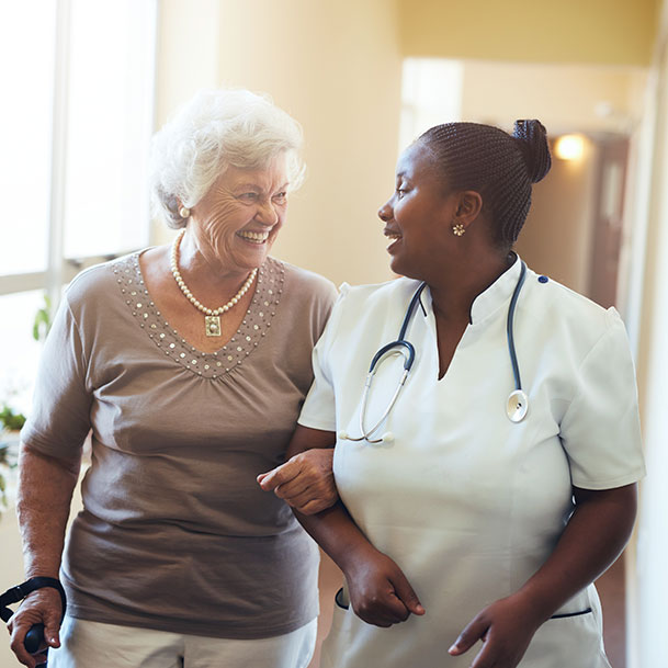 An elderly woman with a cane walking down a corridor with an African American nurse.
