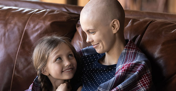 A bald woman sitting on a couch with her arms around a young girl. They are smiling at each other.